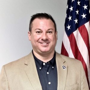 A photo of Samuel Verniero, Jr. posed in front of the American flag.